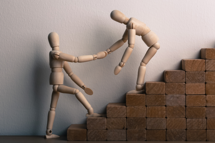 A wooden figurine helping another up a flight of stairs made of wood blocks